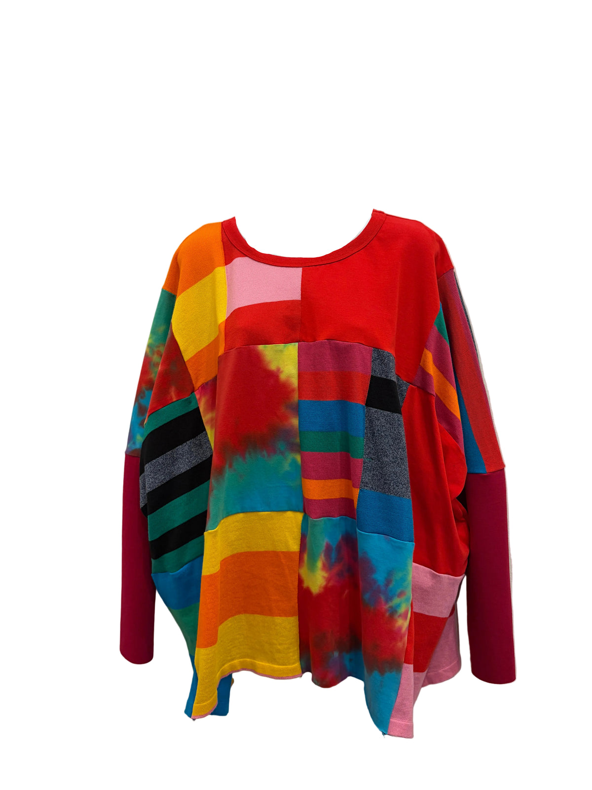 A long-sleeve patchwork top displayed on a white mannequin torso against a plain beige wall. The top features a vibrant mix of solid red, tie-dye patterns, and colorful geometric blocks in pink, yellow, blue, green, and black.
