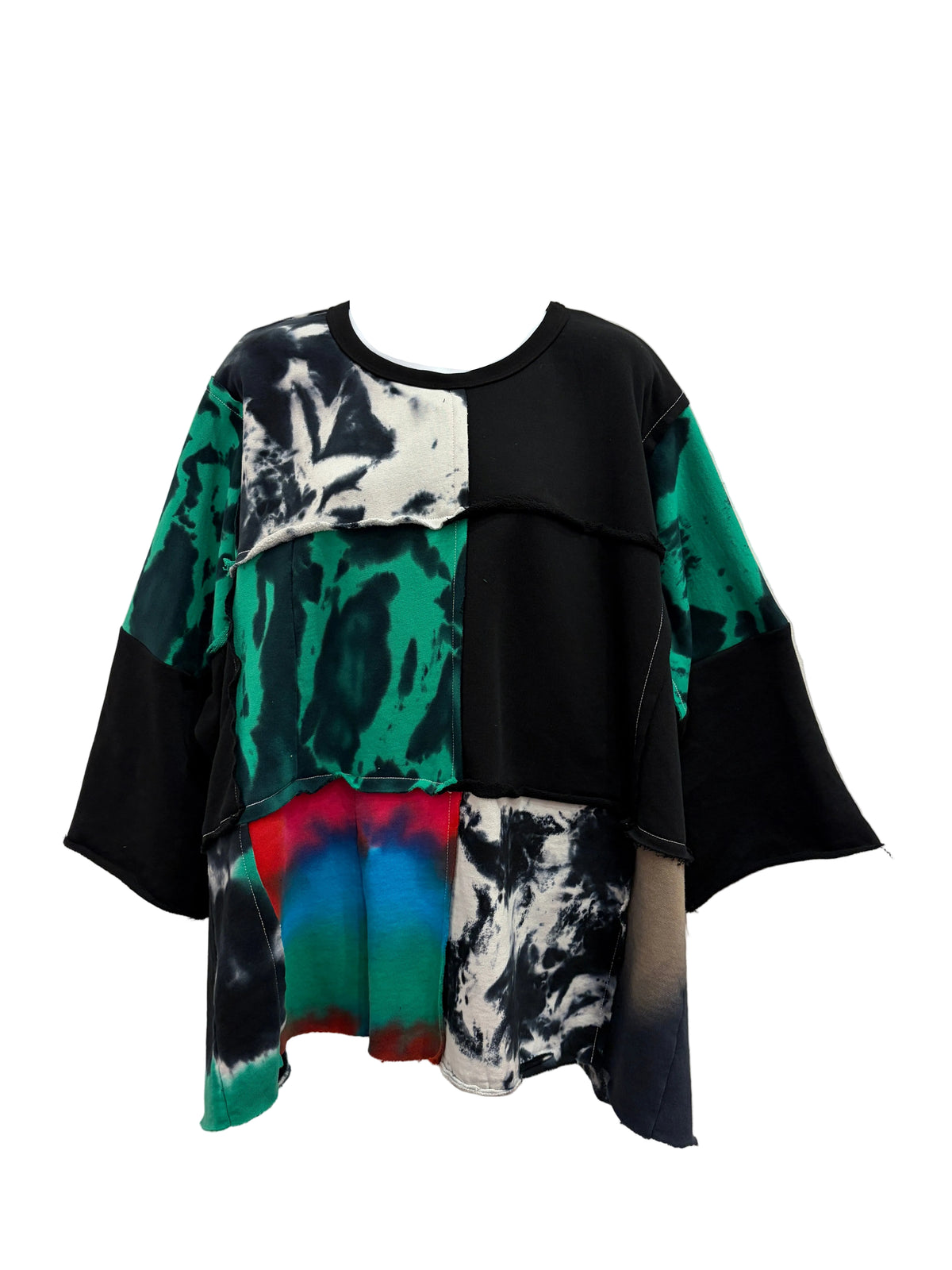 A patchwork short-sleeve top displayed on a white mannequin torso against a light beige wall. The top features a mix of solid black, green and black tie-dye, white and black abstract patterns, and a small red, blue, and green gradient block near the hem.