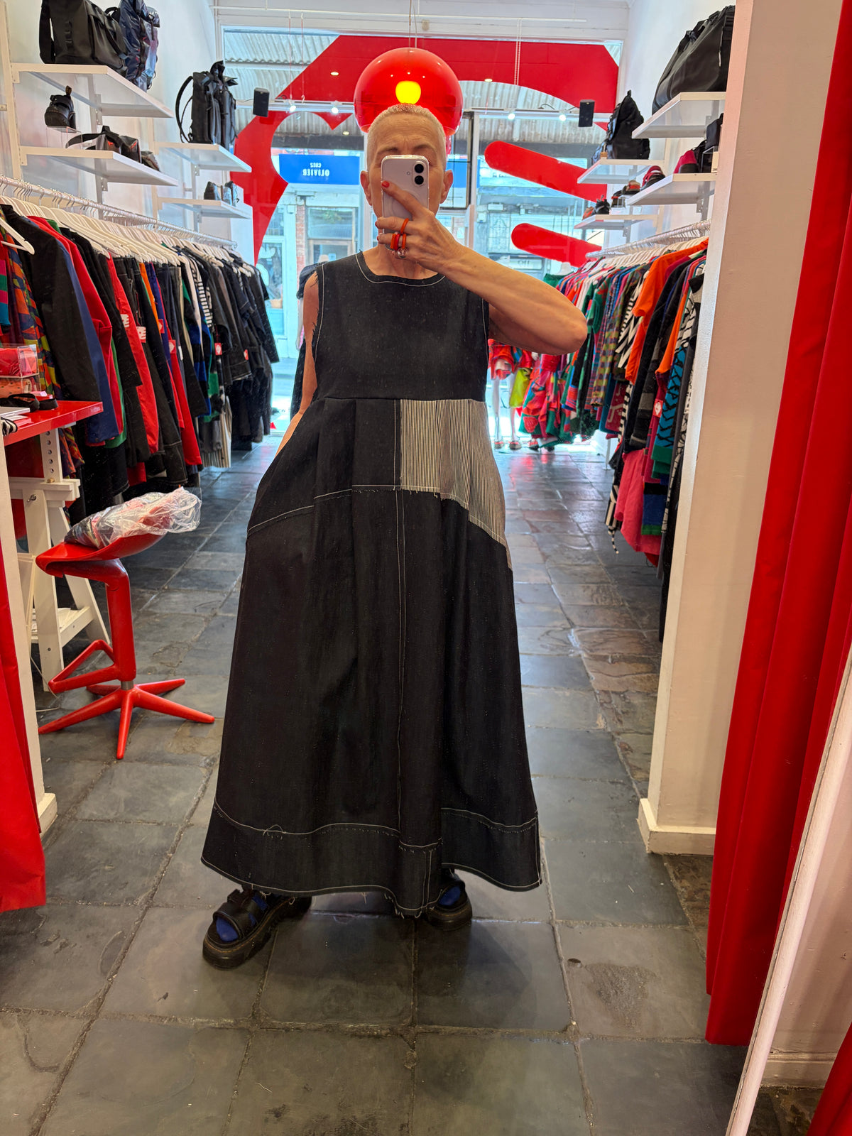 A person taking a mirror selfie in a clothing boutique, wearing a sleeveless dark gray patchwork maxi dress with a wide collar and dark denim-style shoes.