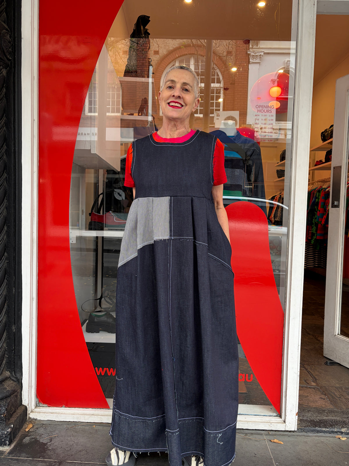 person wearing denim patched dress with white stitching and one stripe white panel at the front. a store front with red graphic is behind them.